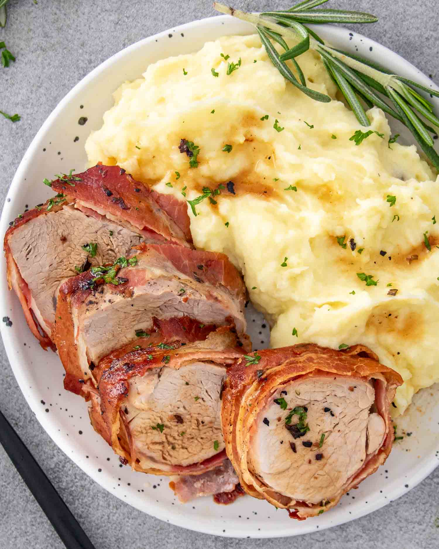 a few sliced of bacon wrapped pork tenderloin on a white plate along some mashed potatoes.