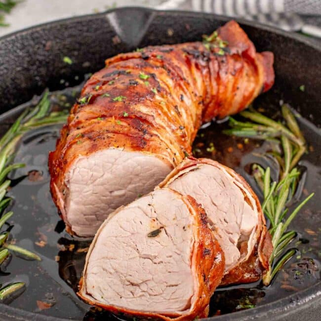 a freshly made bacon wrapped pork tenderloin in a skillet garnished with some fresh rosemary.