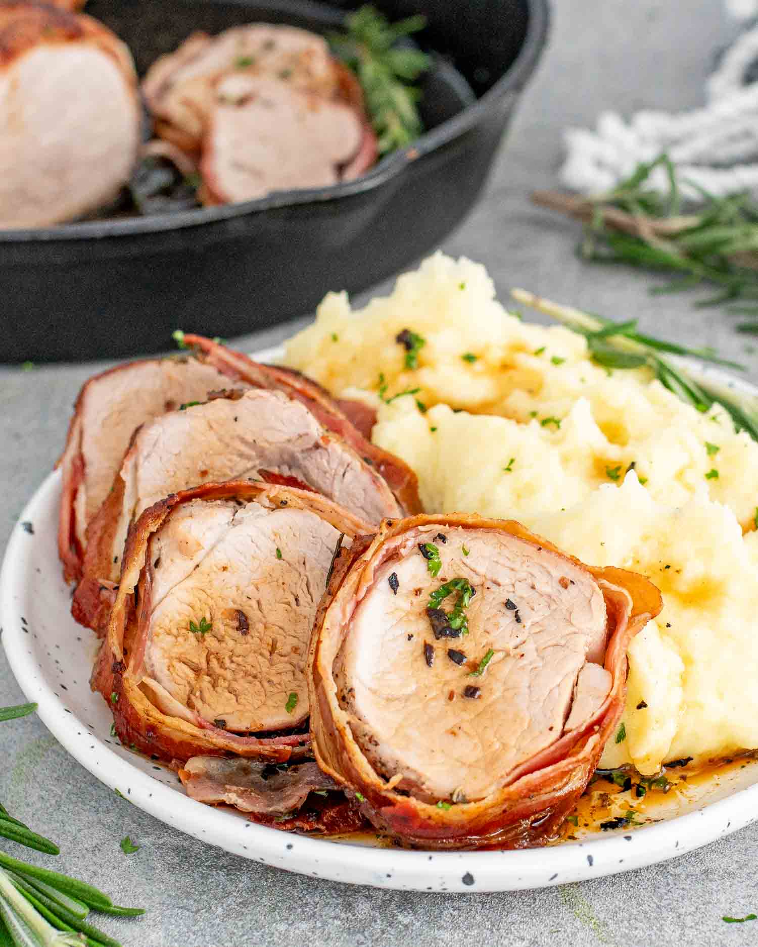 a few sliced of bacon wrapped pork tenderloin on a white plate along some mashed potatoes.