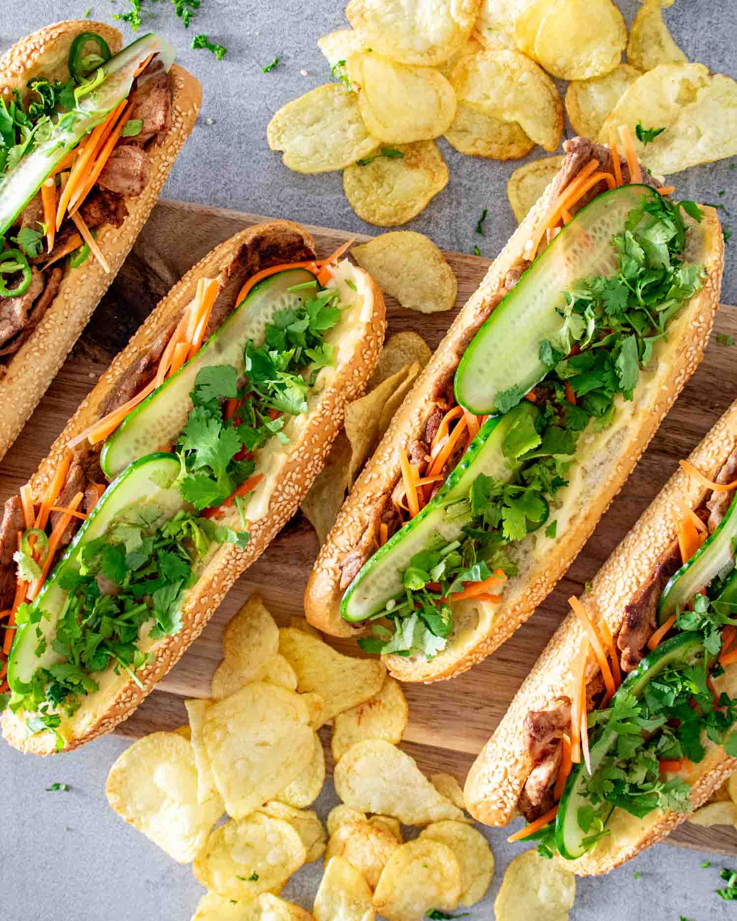 4 banh mi sandwiches on a cutting board with some chips around it.