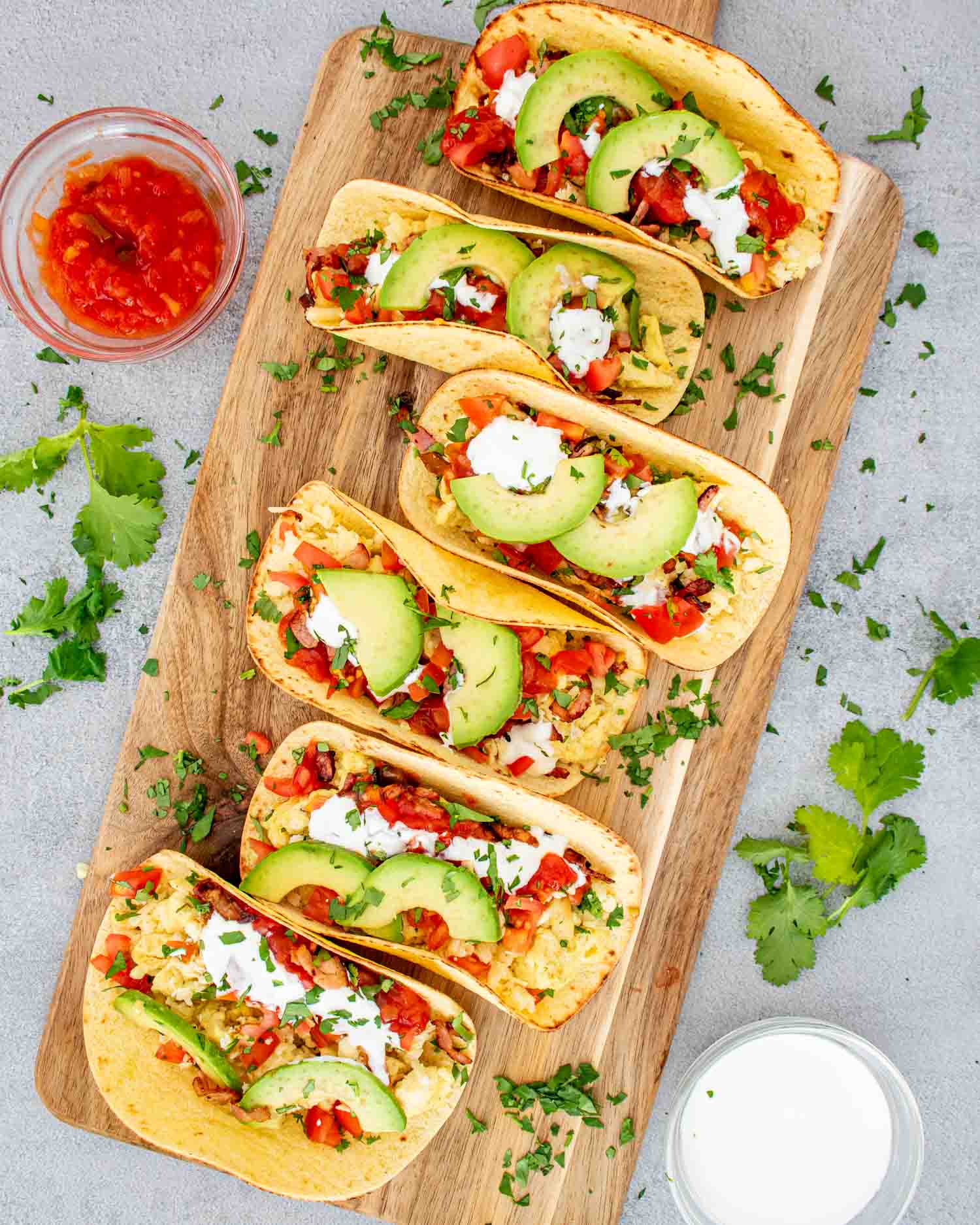 a cutting board with 6 tacos garnished with sour cream, salsa and avocados.