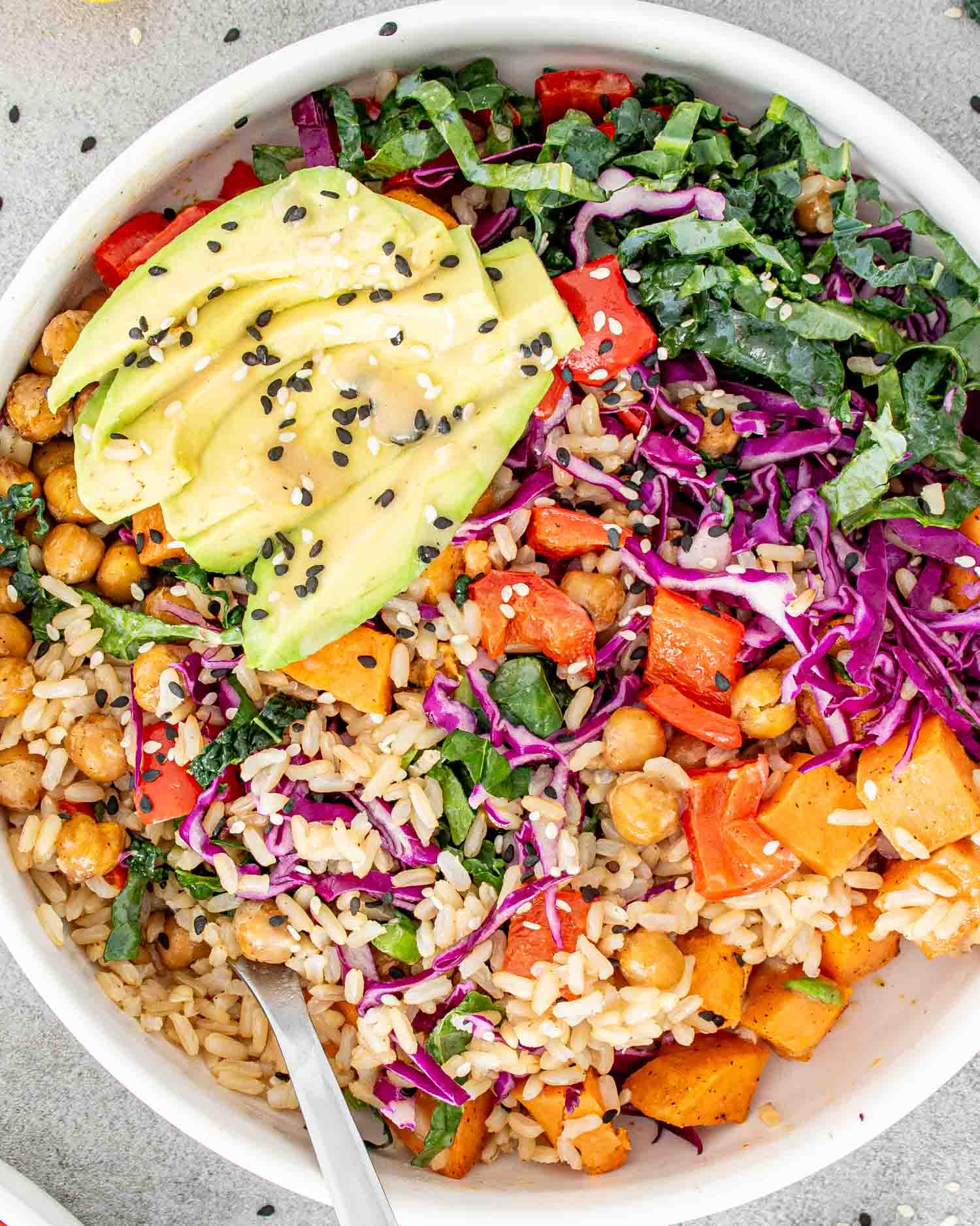 A colorful Buddha bowl with avocado, chickpeas, rice, and mixed vegetables, garnished with black sesame seeds.