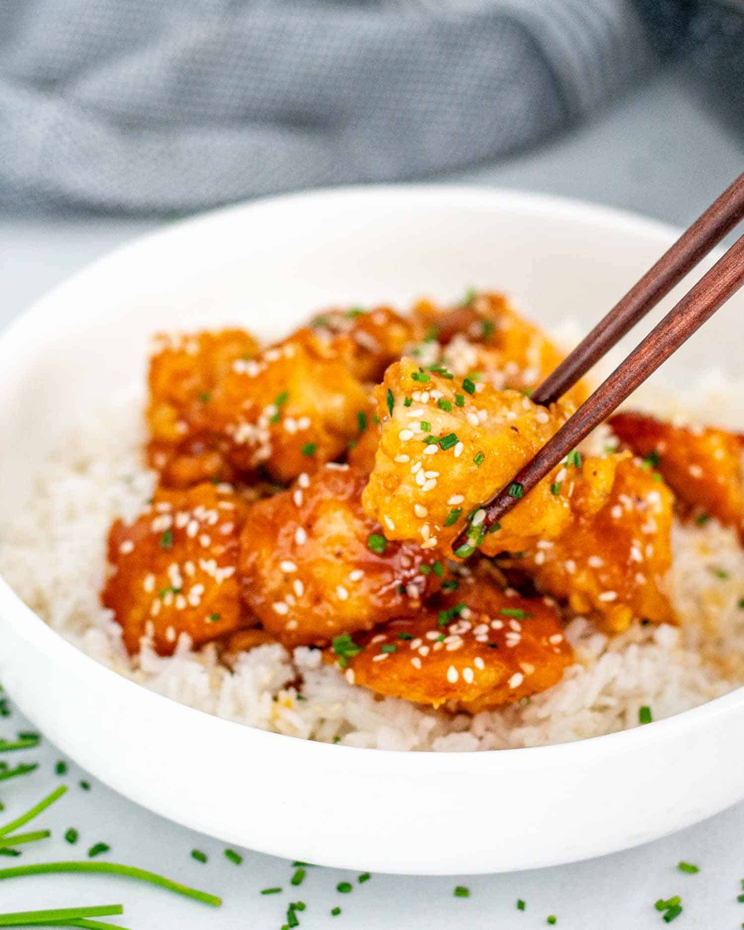 orange chicken on a bed of rice garnished with chives in a white bowl.