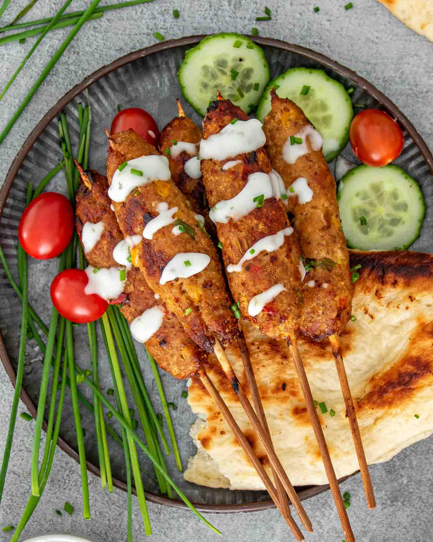 chicken kebabs drizzled with tzatziki sauce on a plate alongside some pita bread and cucumbers.