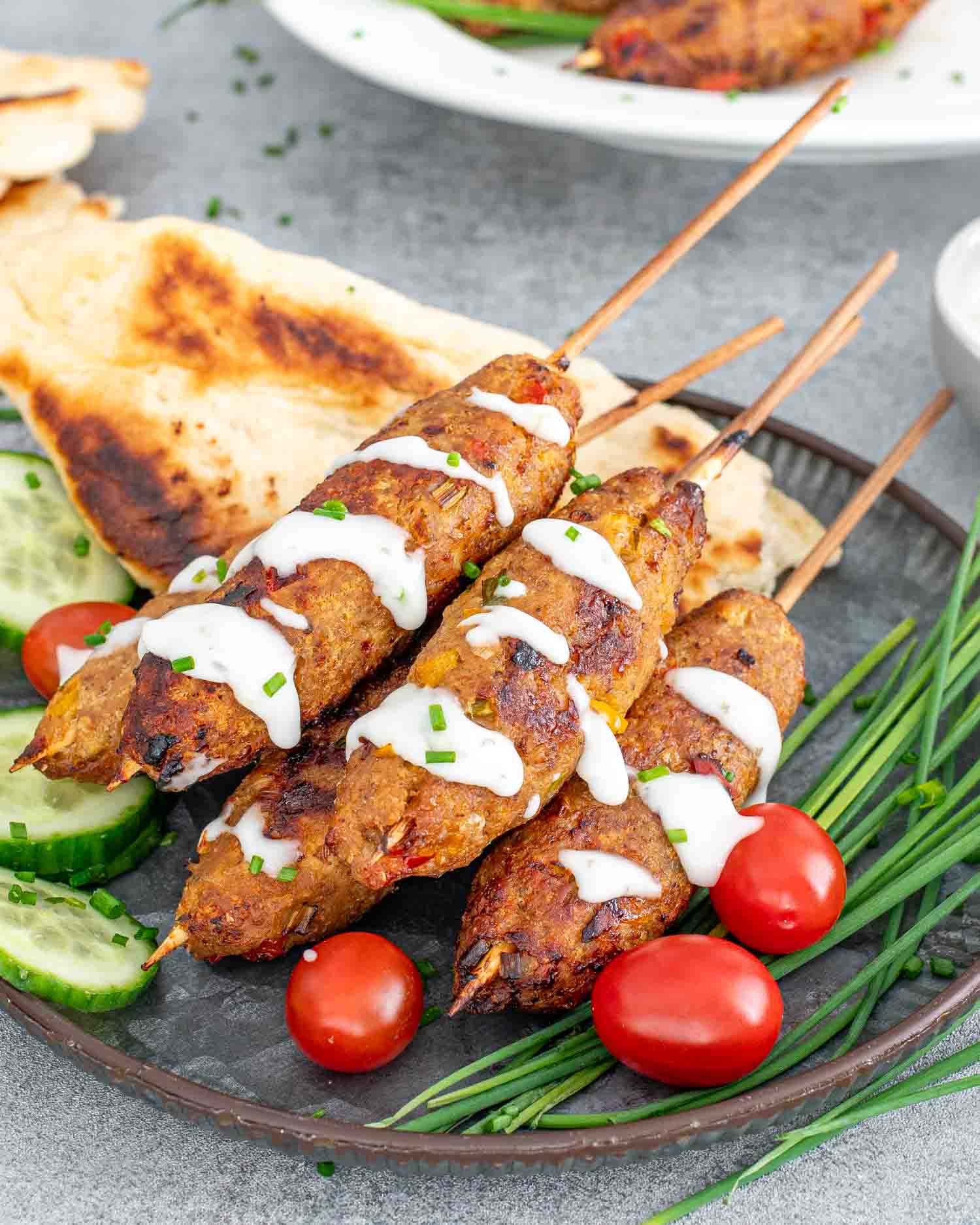 chicken kebabs drizzled with tzatziki sauce on a plate alongside some pita bread and cucumbers.