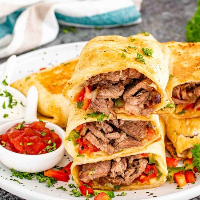 a steak of cheesesteak burritos on a white plate with salsa and sour cream.