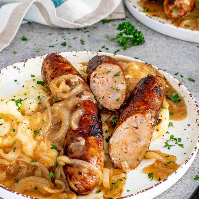 a serving of bangers and mash with onion gravy on a white plate garnished with parsley.