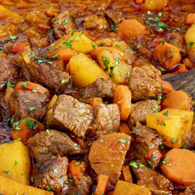 A close-up shot of a hearty Beef Goulash in a large Dutch oven. Tender chunks of beef, golden potatoes, and sliced carrots are simmered in a rich, deep-red paprika-infused broth. The dish is garnished with freshly chopped parsley, adding a pop of green.