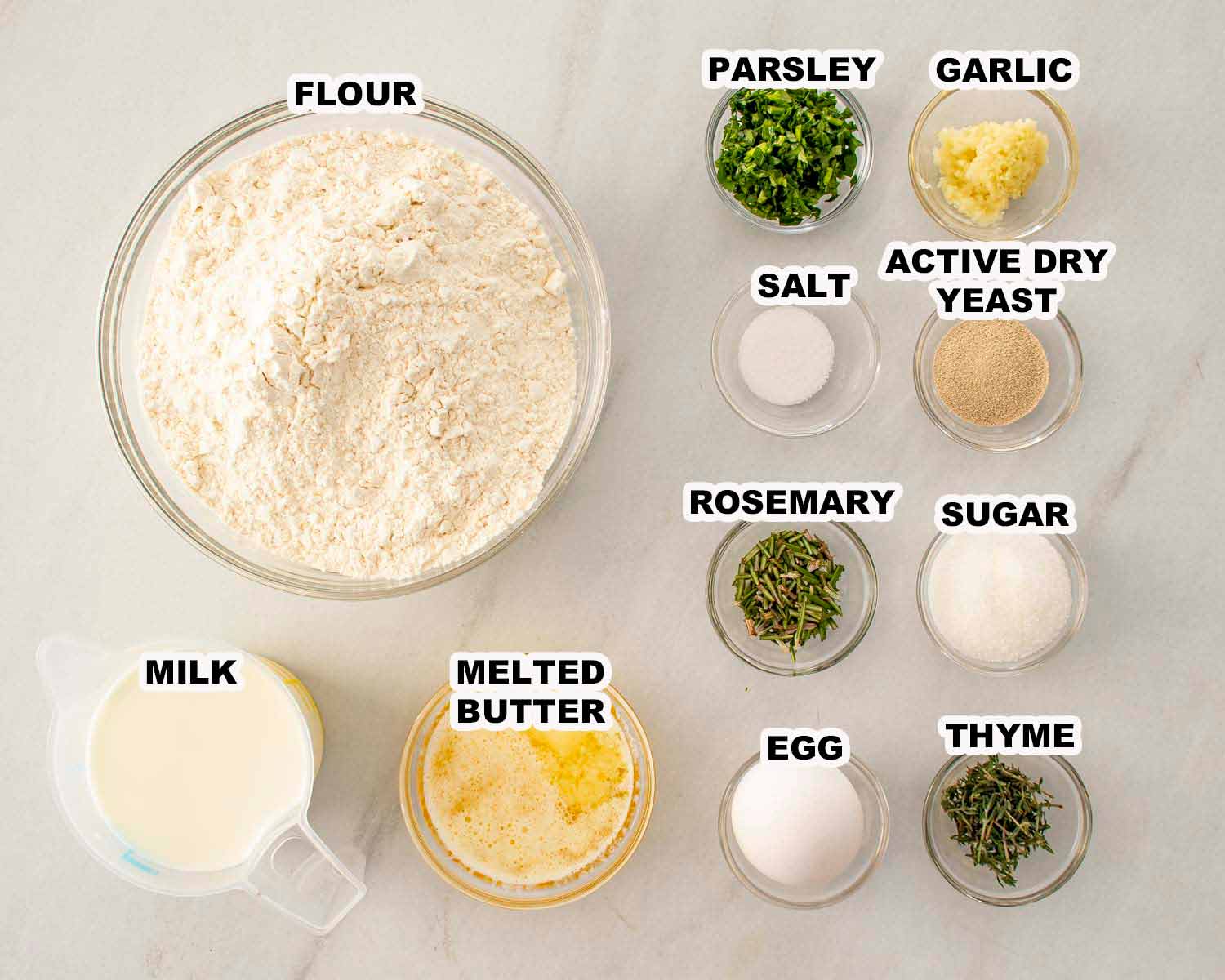 Ingredients for garlic herb pull apart rolls arranged in bowls, including flour, yeast, milk, butter, garlic, and fresh herbs.