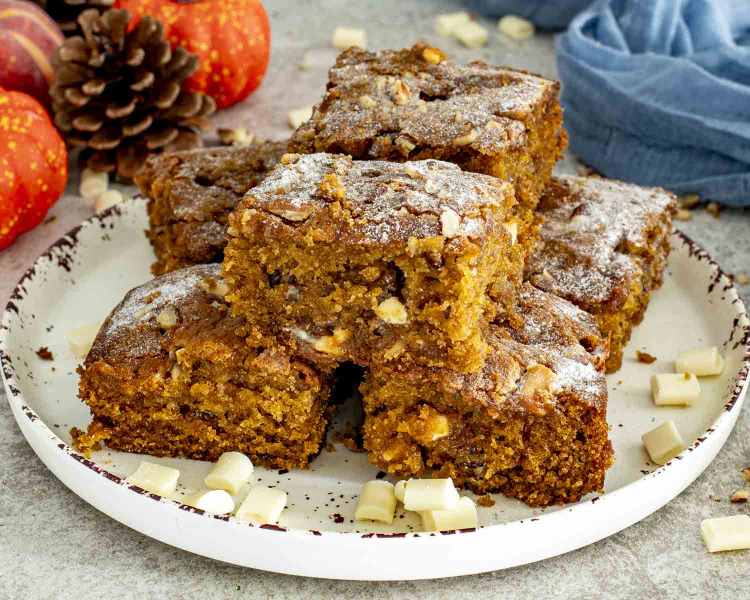 Stack of pumpkin spice blondies on a serving plate, studded with white chocolate and nuts, lightly dusted with powdered sugar.