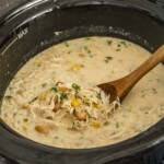 Creamy slow cooker white chicken chili simmering with shredded chicken, beans and corn, stirred gently with a wooden spoon.