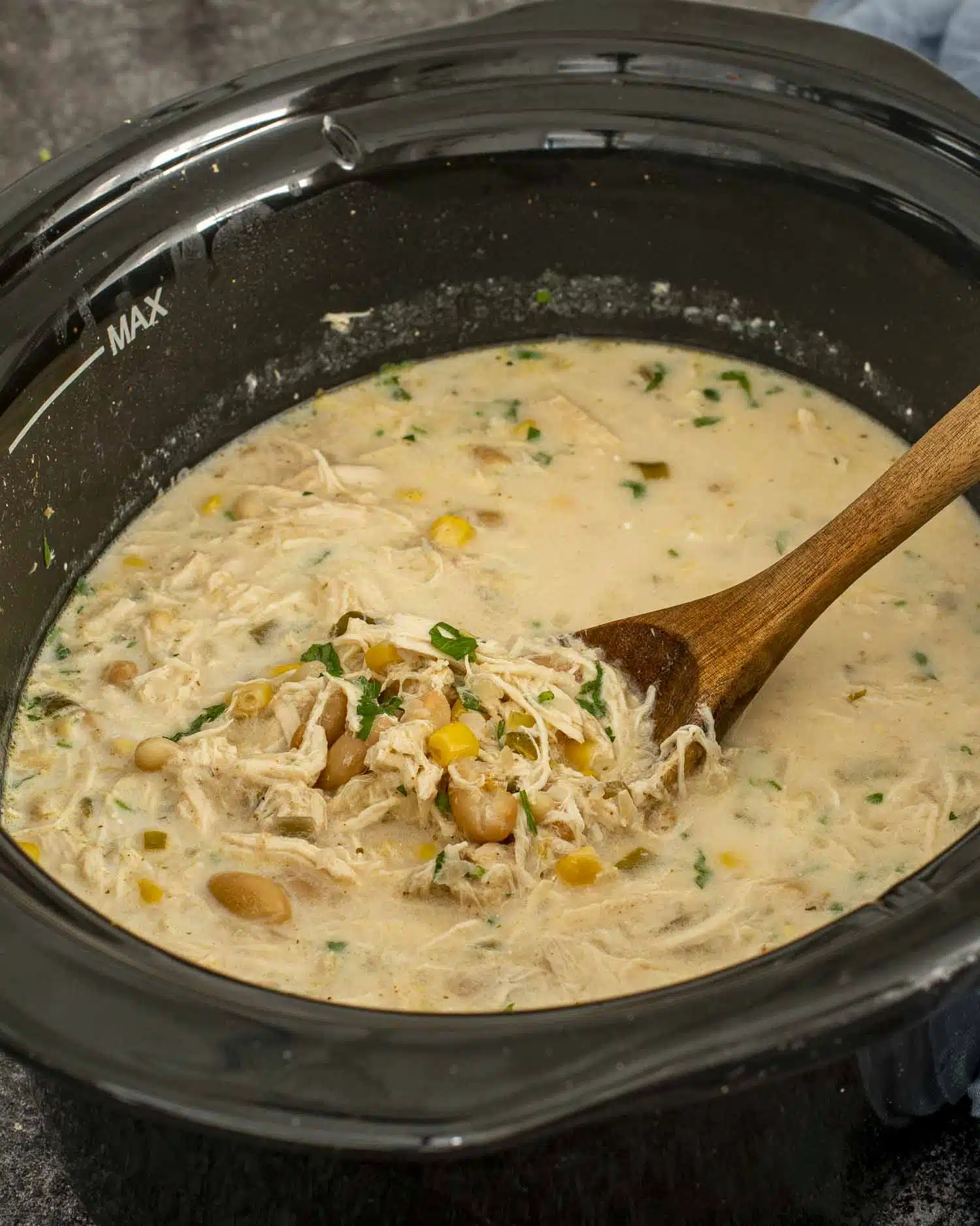 Creamy slow cooker white chicken chili simmering with shredded chicken, beans and corn, stirred gently with a wooden spoon.