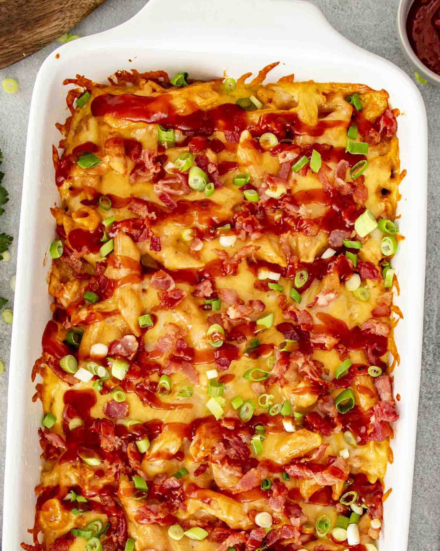 Overhead view of BBQ chicken casserole topped with melted cheddar, crispy bacon, green onions, and barbecue sauce in baking dish.