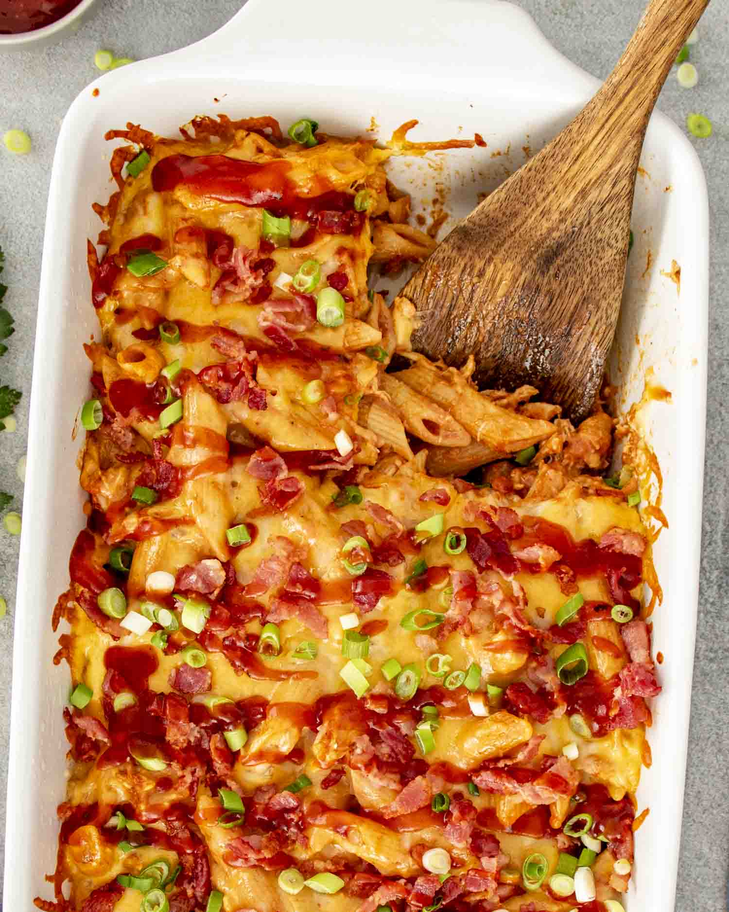 Spoon serving BBQ chicken casserole showing cheesy penne pasta, shredded chicken, bacon bits, green onions, and barbecue sauce.