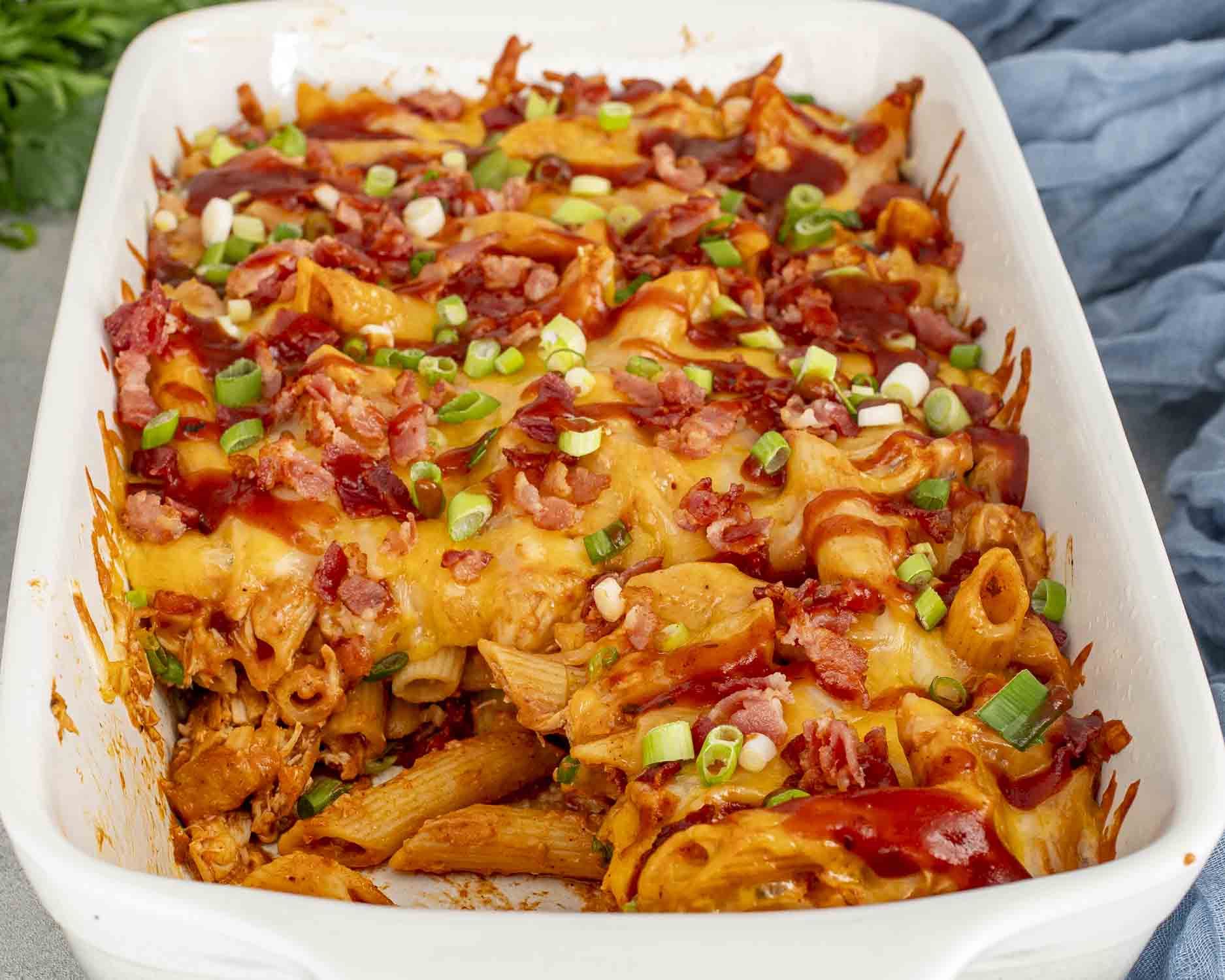 BBQ chicken casserole baked in a white casserole dish with penne pasta, melted cheese, bacon, green onions, and barbecue sauce.