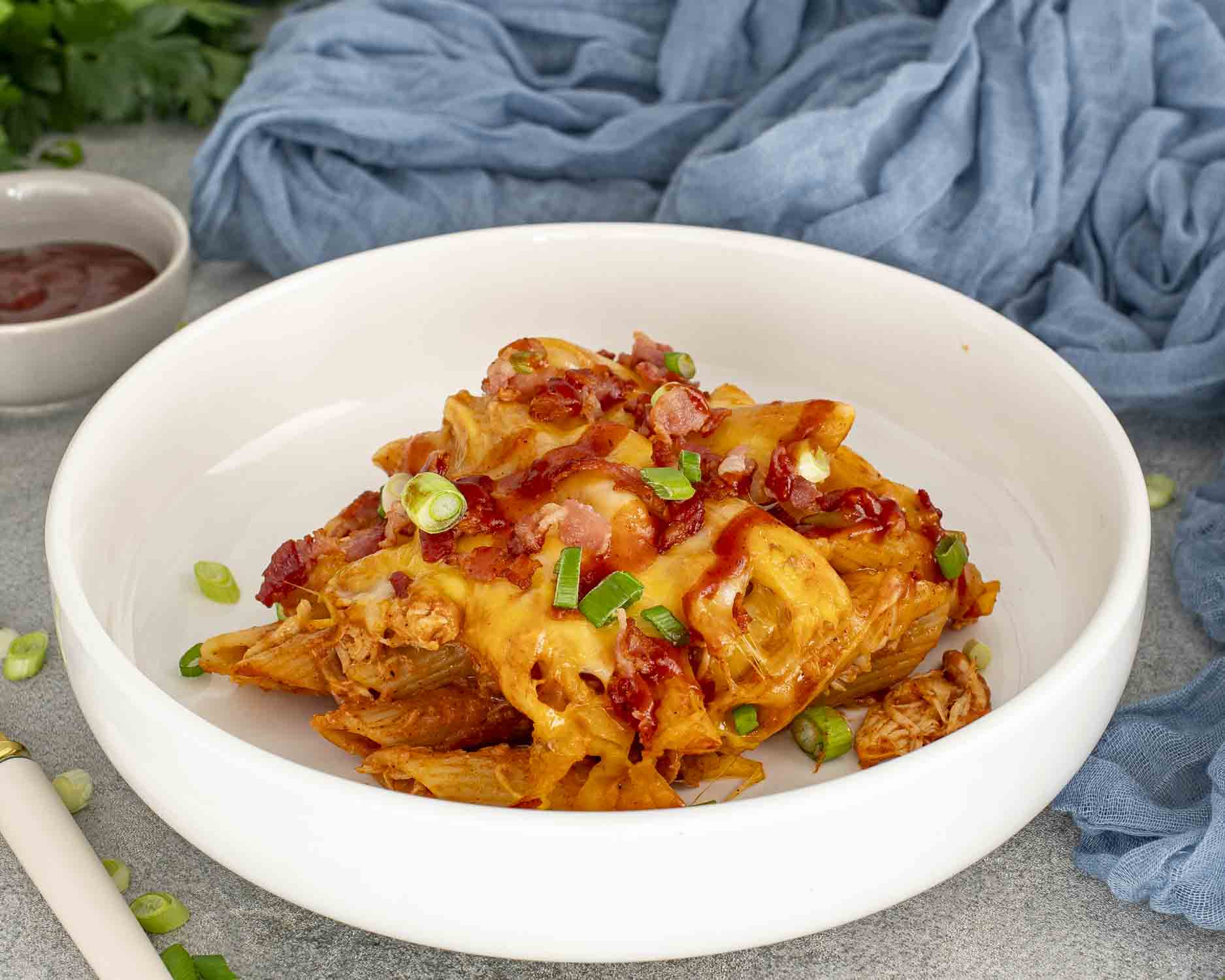 Plated BBQ chicken casserole with penne pasta, melted cheddar cheese, crispy bacon, green onions, and smoky barbecue sauce drizzle.