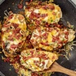 Smothered chicken breasts cooking in a skillet with melted cheese, crispy bacon, sautéed mushrooms, and caramelized onions.