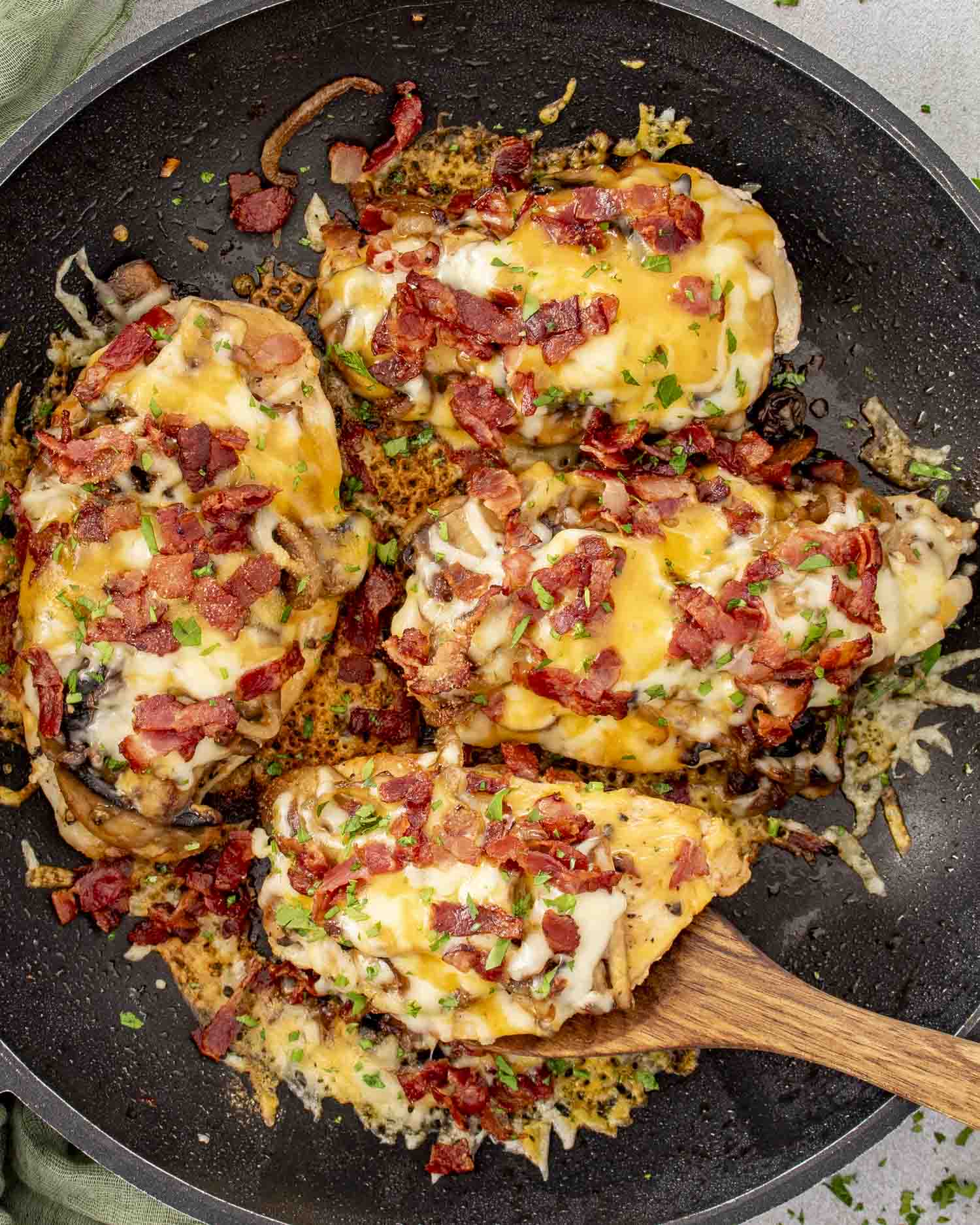 Smothered chicken breasts cooking in a skillet with melted cheese, crispy bacon, saut&eacute;ed mushrooms, and caramelized onions.
