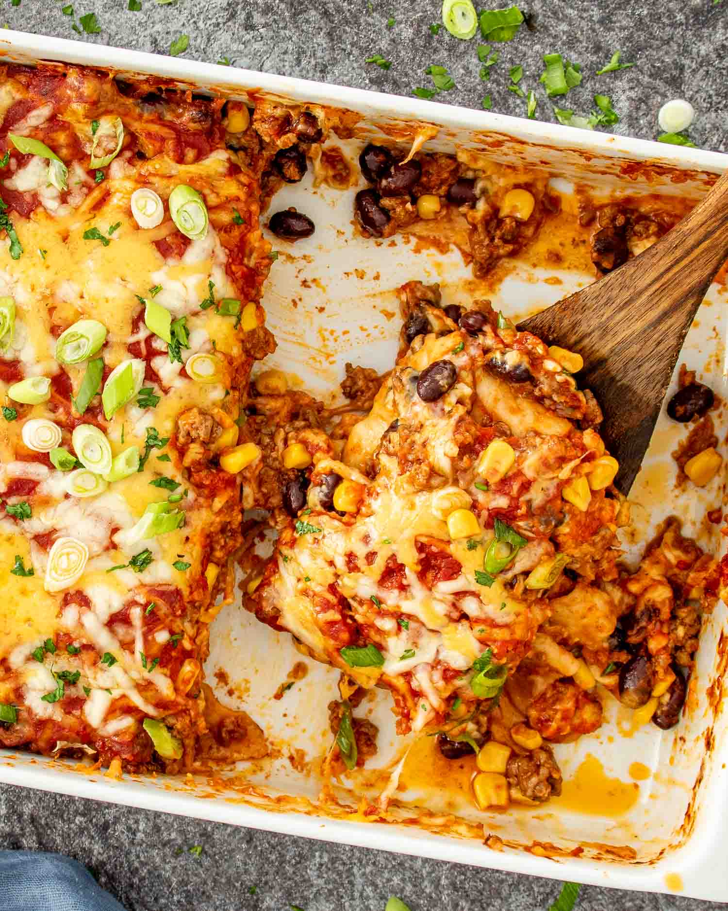 Beef enchilada casserole in a baking dish with a serving spoon lifting a cheesy, saucy portion.