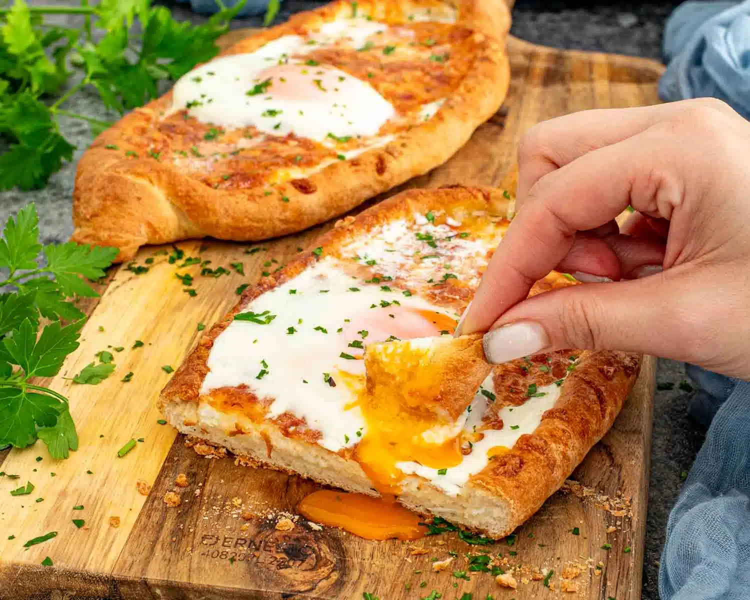 Hand tearing crusty Khachapuri bread and dipping into creamy melted cheese and runny egg yolk.