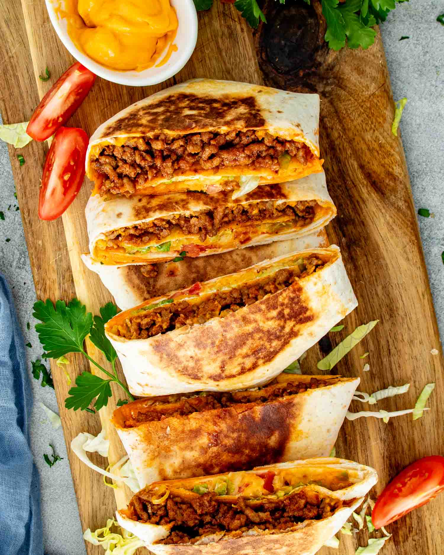 Pan-toasted Taco Bell Crunchwrap Supreme copycat arranged on cutting board with tomato slices and nacho cheese sauce nearby.