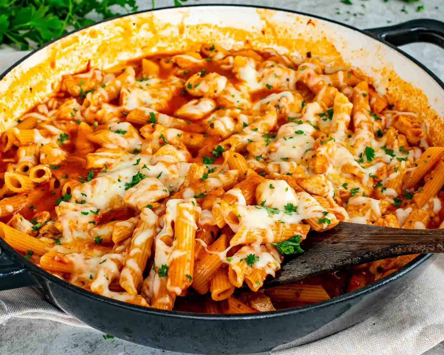 Chicken parmesan pasta skillet in large pan with penne, tender chicken, marinara sauce, and melted mozzarella being scooped with spoon.