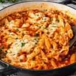 Chicken parmesan pasta skillet in large pan with penne, tender chicken, marinara sauce, and melted mozzarella being scooped with spoon.