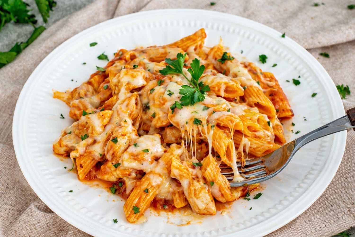 Serving of chicken parmesan pasta skillet on white plate with penne, juicy chicken, marinara sauce, melted cheese, and parsley garnish.