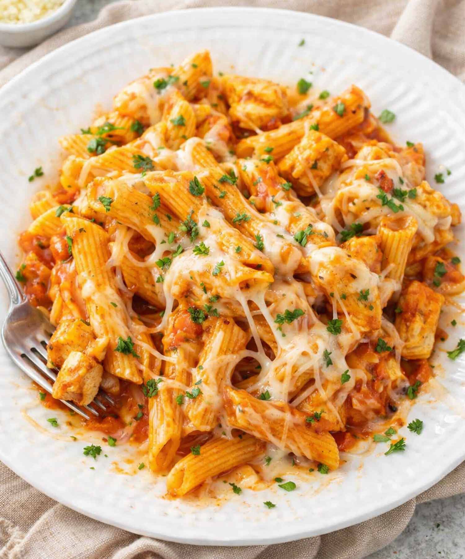 Serving of chicken parmesan pasta skillet on white plate with penne, juicy chicken, marinara sauce, melted cheese, and parsley garnish.