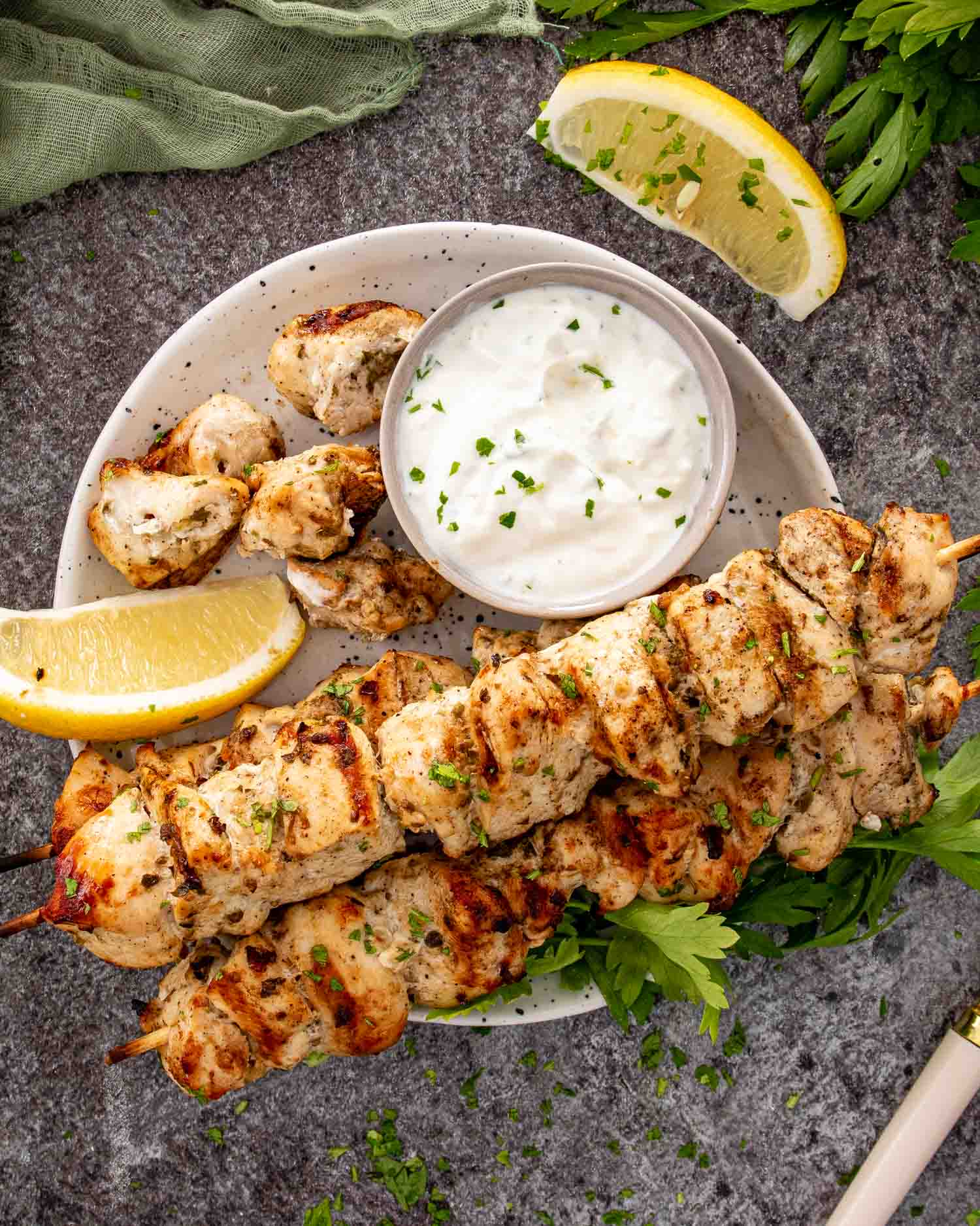 Top view grilled chicken skewers arranged on white platter with lemon wedges, parsley garnish, and gray stone background.