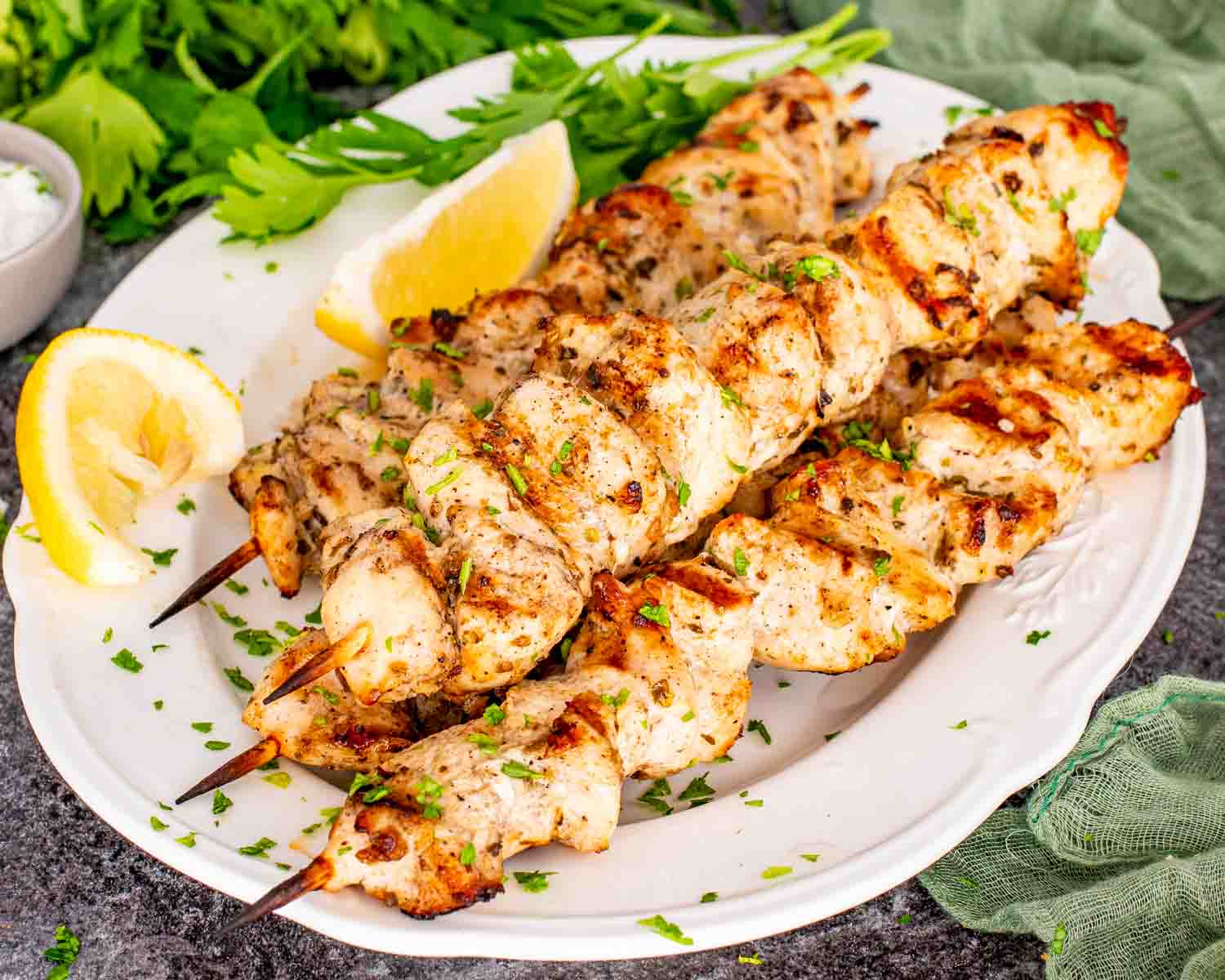 Overhead view grilled chicken skewers on plate with creamy dipping sauce, lemon wedges, parsley, and rustic stone background.