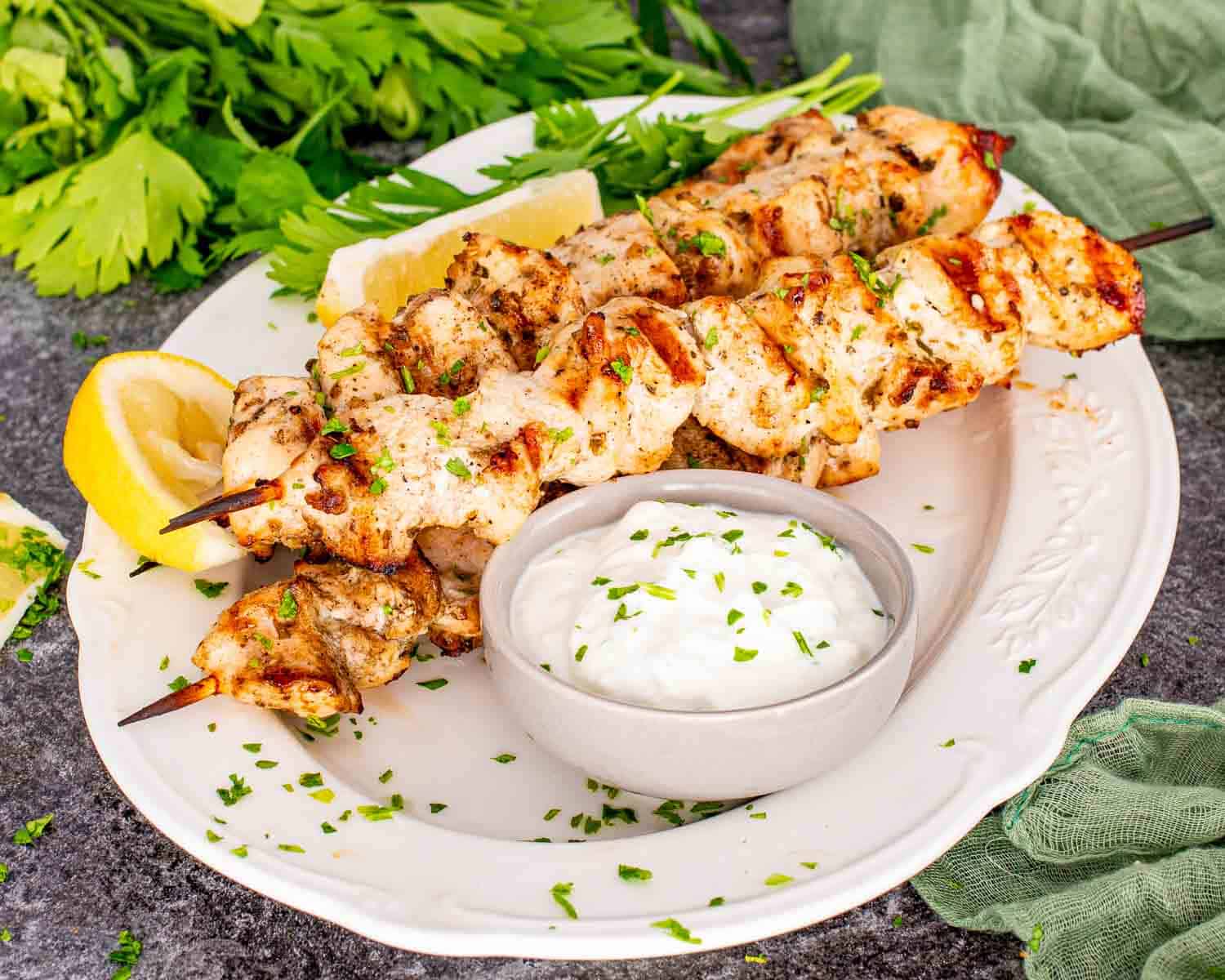 Grilled lemon garlic chicken skewers on oval white platter with parsley, lemon wedges, and textured green cloth on stone surface.