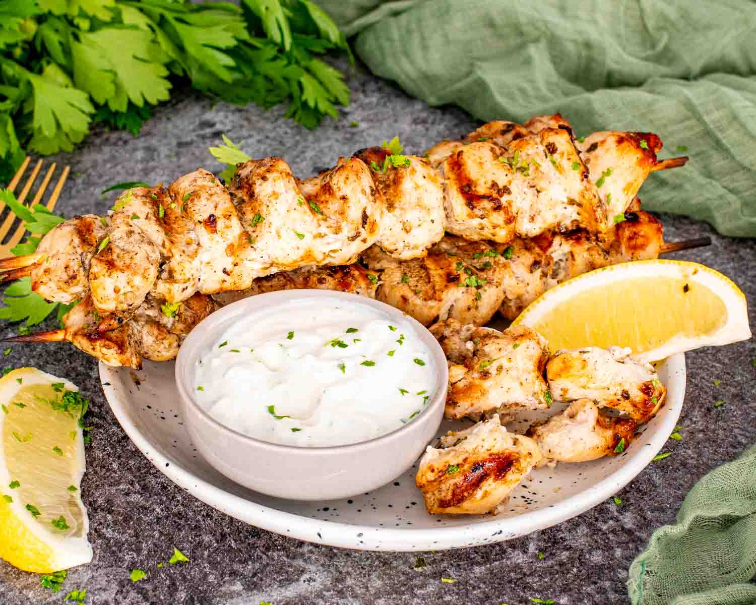 Grilled lemon garlic chicken skewers on oval white platter with parsley, lemon wedges, and textured green cloth on stone surface.