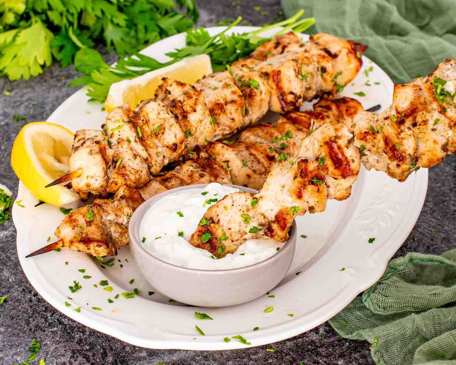 Grilled chicken skewers stacked on plate with creamy white dipping sauce, parsley garnish, lemon wedge, and green cloth napkin.