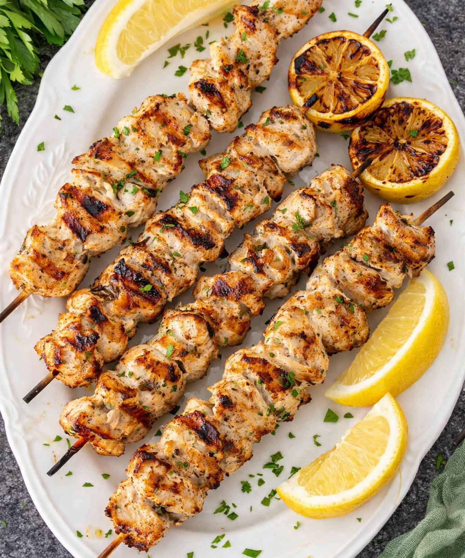 Grilled lemon garlic chicken skewers on white platter with charred lemon halves, parsley, and lemon wedges on stone surface.