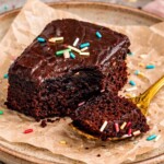 Fudgy lunch lady brownie with smooth chocolate frosting, rich moist crumb visible, plated with parchment and colorful sprinkles scattered around.