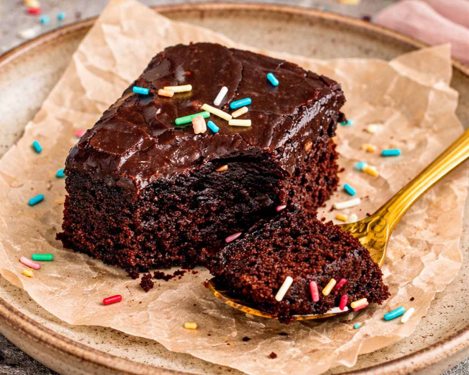 Fudgy lunch lady brownie with smooth chocolate frosting, rich moist crumb visible, plated with parchment and colorful sprinkles scattered around.