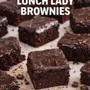 Classic lunch lady brownies cut into squares with shiny chocolate frosting, rich fudgy texture visible, scattered chocolate chunks on parchment background.Classic lunch lady brownies cut into squares with shiny chocolate frosting, rich fudgy texture visible, scattered chocolate chunks on parchment background.