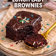 Soft fudgy lunch lady brownies with glossy chocolate frosting, bite taken, served on parchment with colorful sprinkles and gold spoon.