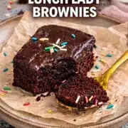 Soft fudgy lunch lady brownies with glossy chocolate frosting, bite taken, served on parchment with colorful sprinkles and gold spoon.