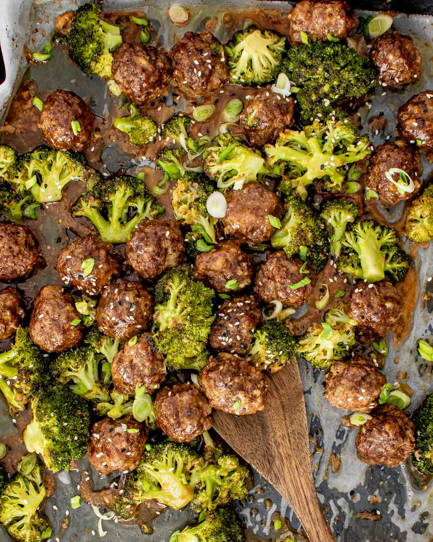 Wooden spoon lifting a glossy honey garlic meatball from a parchment lined sheet pan with roasted broccoli.