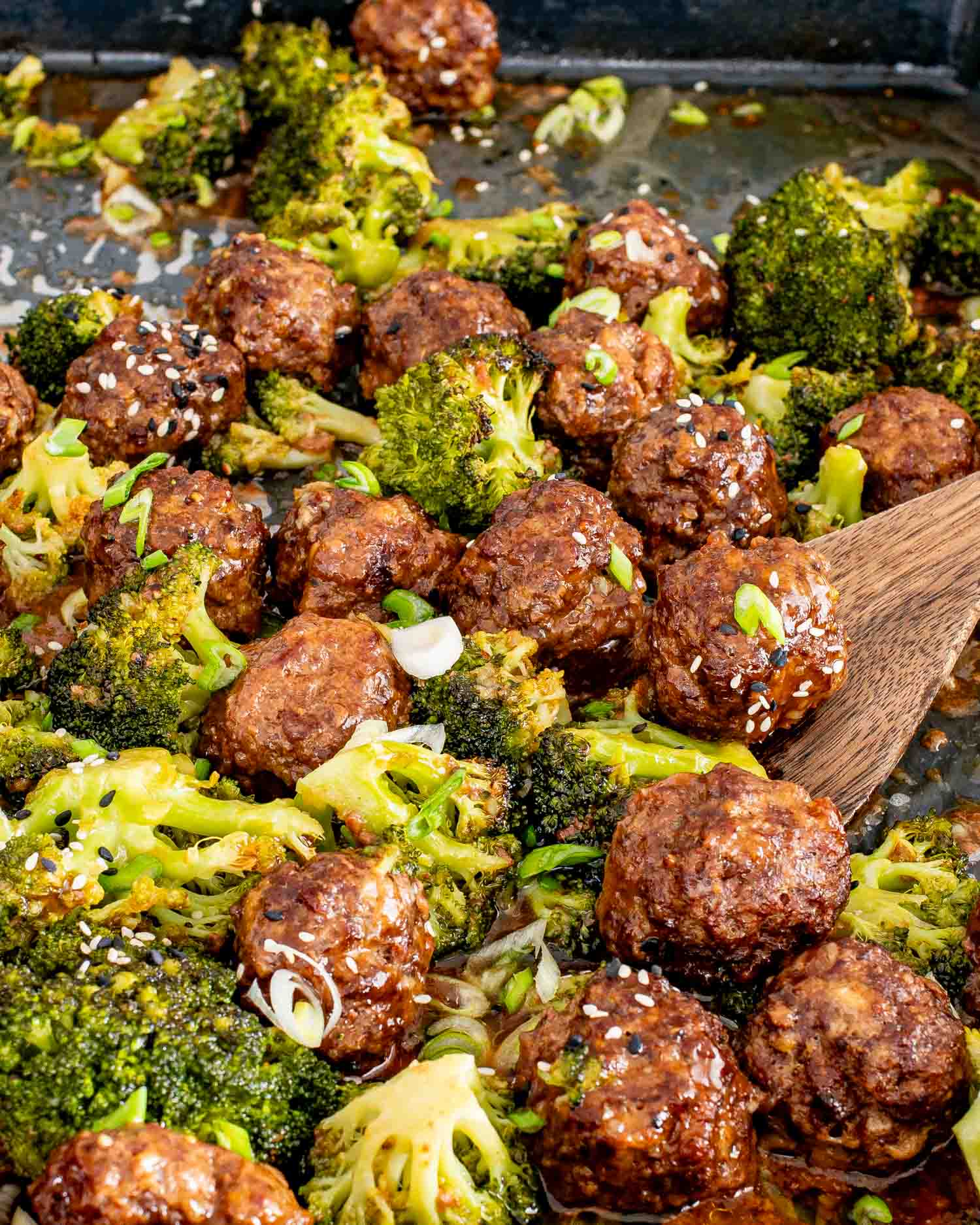 Wooden spoon lifting a glossy honey garlic meatball from a parchment lined sheet pan with roasted broccoli.