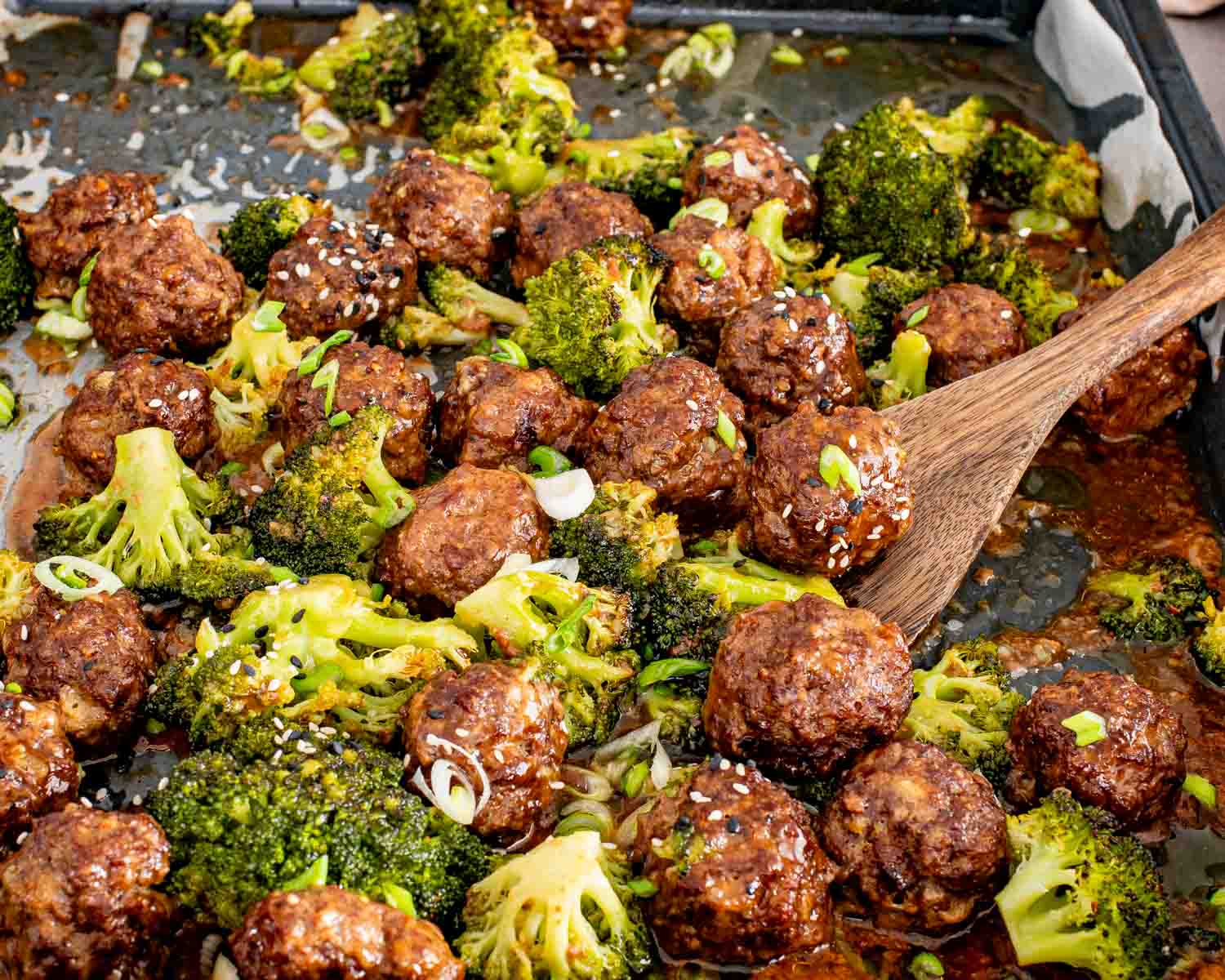 Wooden spoon lifting a glossy honey garlic meatball from a parchment lined sheet pan with roasted broccoli.