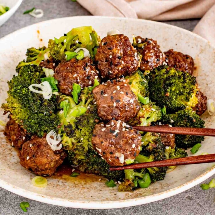 Close up of glazed honey garlic meatballs and broccoli in a white bowl with wooden chopsticks.