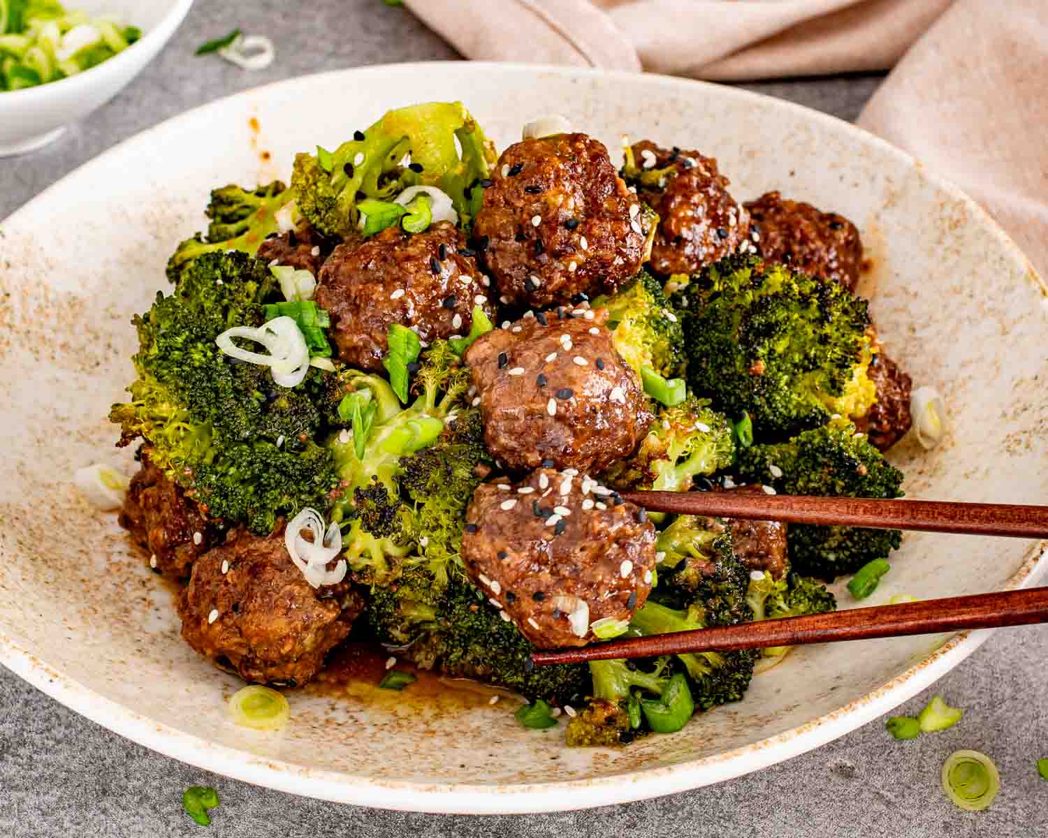 Close up of glazed honey garlic meatballs and broccoli in a white bowl with wooden chopsticks.