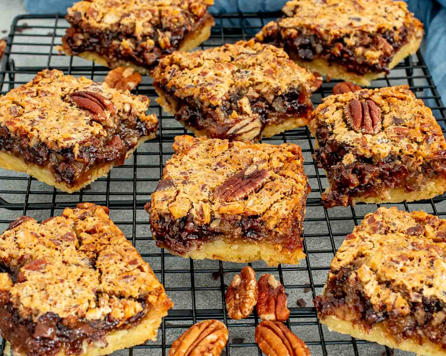 Overhead view of nine chocolate chip pecan pie bars cooling on a black wire rack, topped with whole pecans and showing the caramelized pecan and chocolate filling, with whole pecans scattered below.