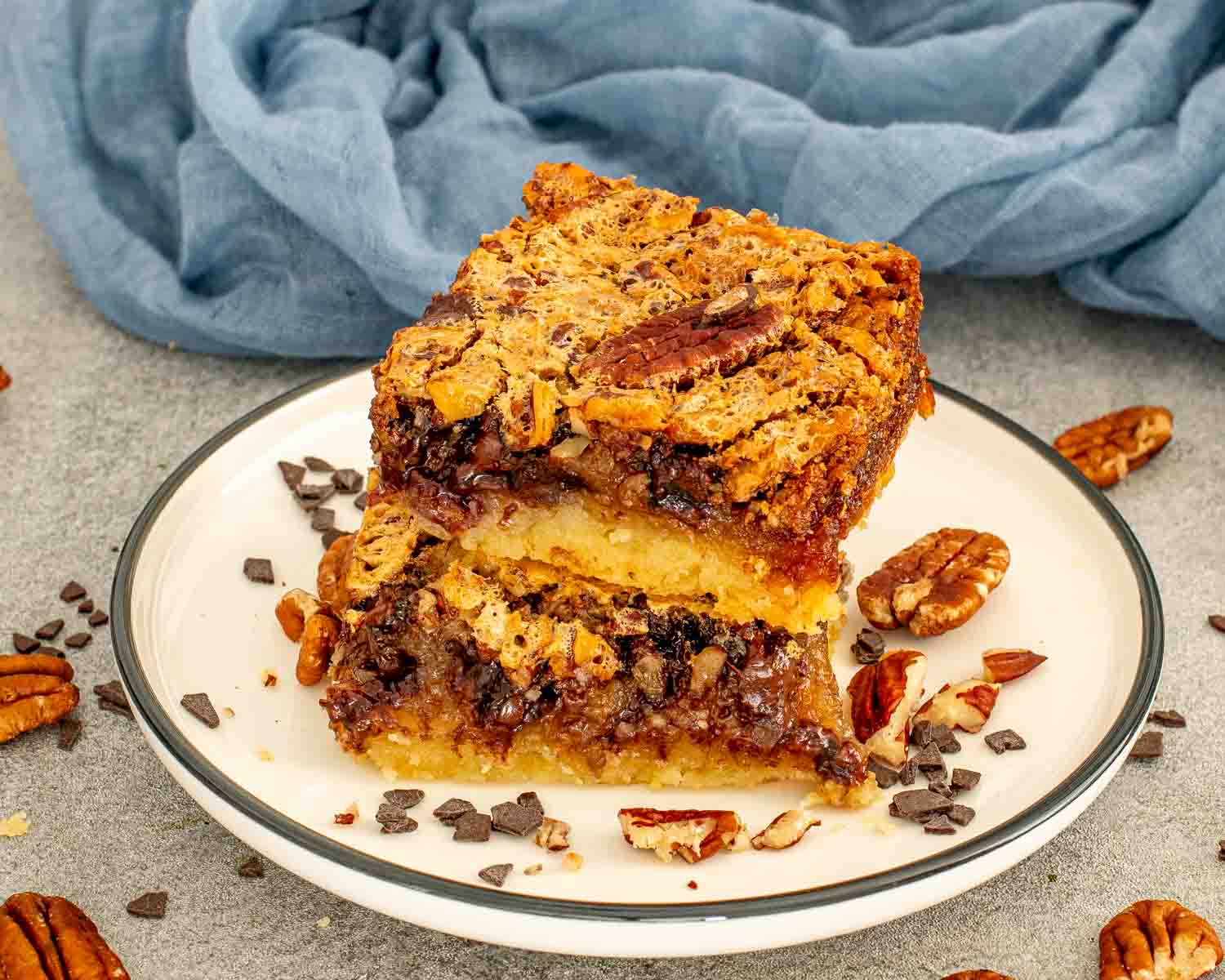 Two chocolate chip pecan pie bars on a white plate with a dark rim, showing the gooey caramel pecan chocolate filling and buttery shortbread base in cross section, with whole pecans and chocolate chunks around the plate.