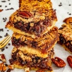 Three chocolate chip pecan pie bars stacked on parchment paper showing the distinct shortbread base and gooey pecan chocolate filling, with scattered pecans, chocolate chips, and a gold fork alongside.