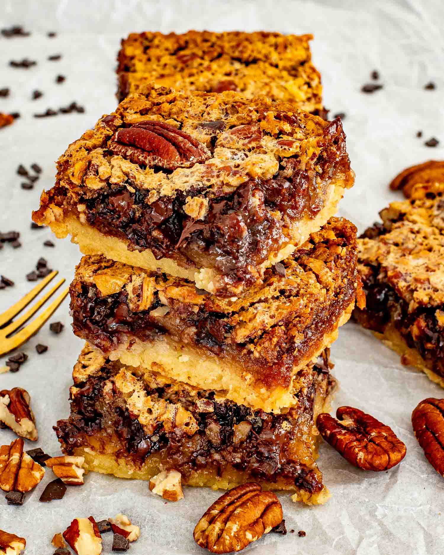 Three chocolate chip pecan pie bars stacked on parchment paper showing the distinct shortbread base and gooey pecan chocolate filling, with scattered pecans, chocolate chips, and a gold fork alongside.