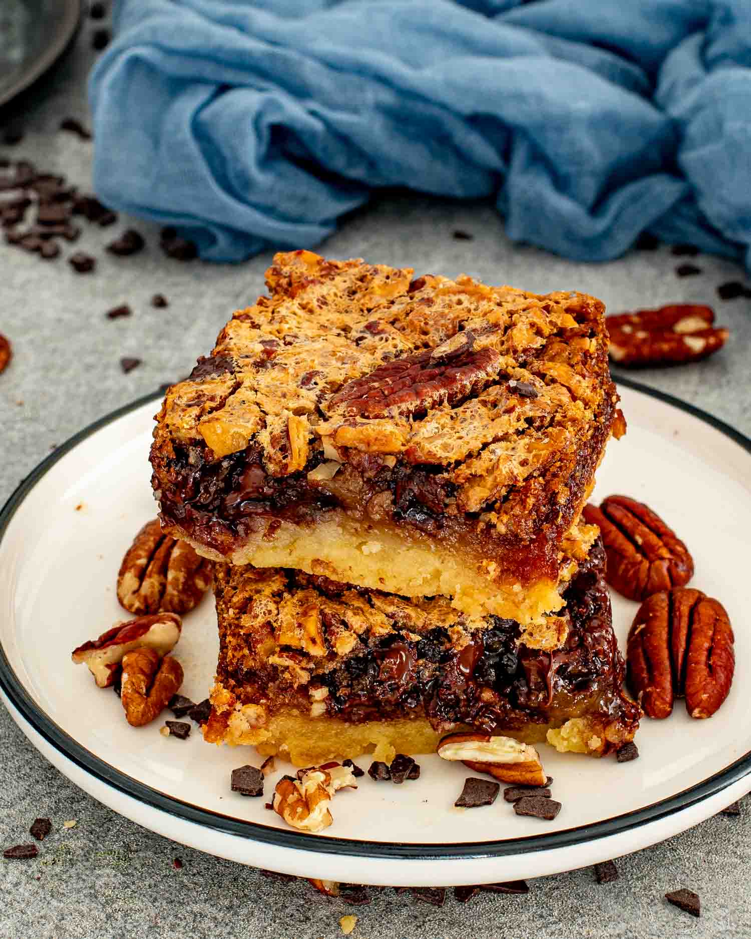 Two chocolate chip pecan pie bars stacked on a white plate with a dark rim, surrounded by whole pecans and chopped chocolate pieces, with a blue linen in the background.